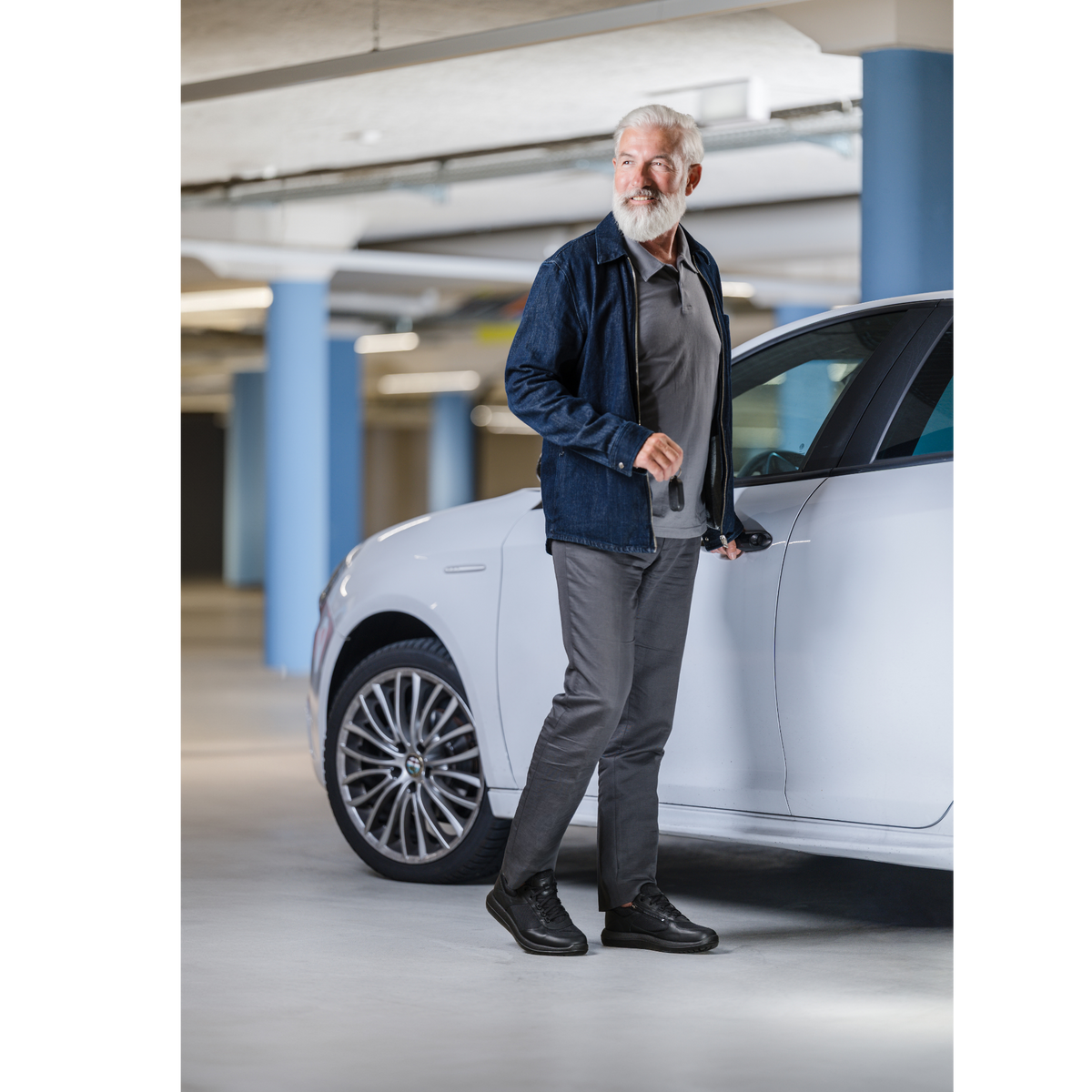 An older man with a white beard stands in a modern parking garage next to a white car, wearing a dark blue denim jacket, gray polo shirt, gray trousers, and black shoes.