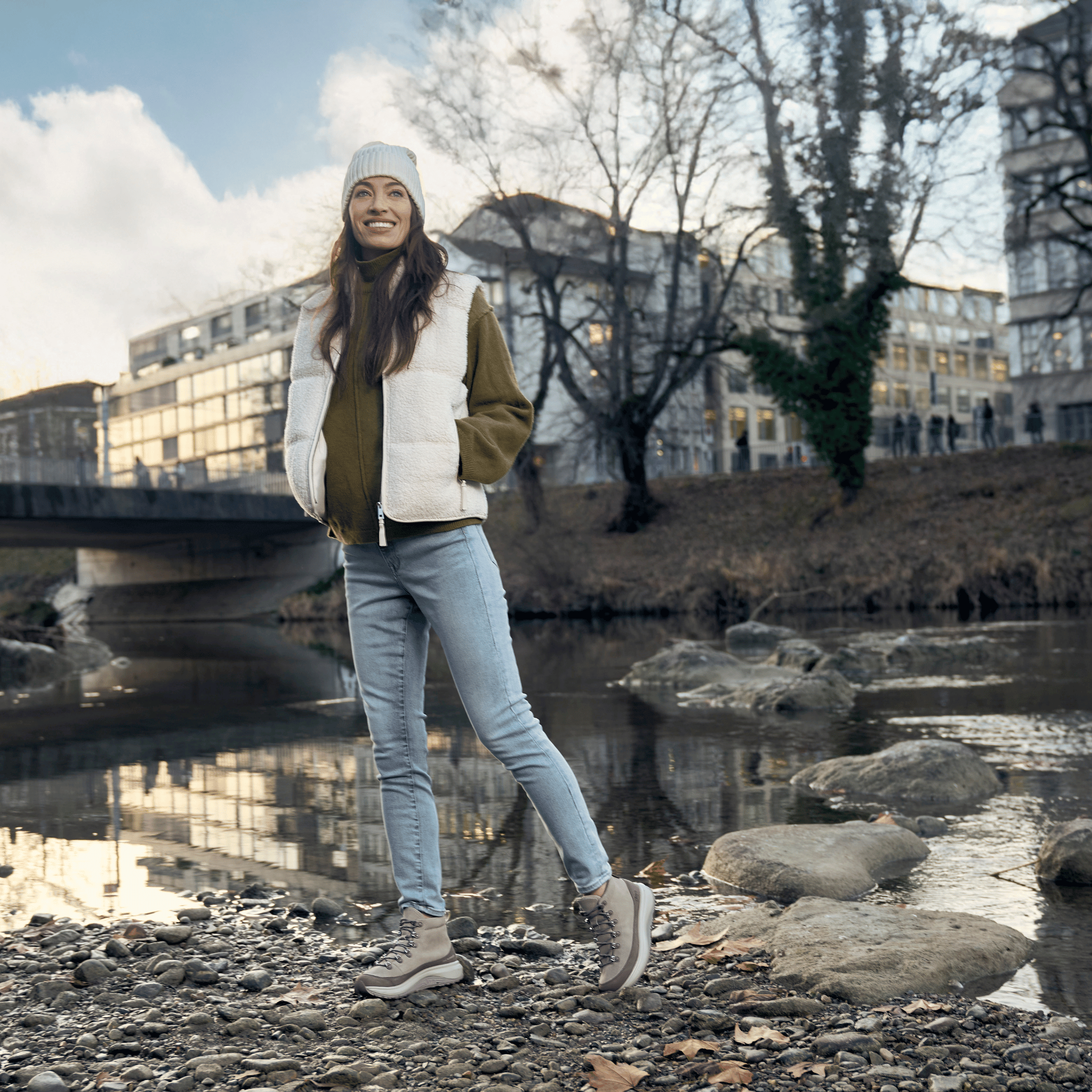women standing on a river bank with buildings and trees blurred in the background wearing jeans and boots