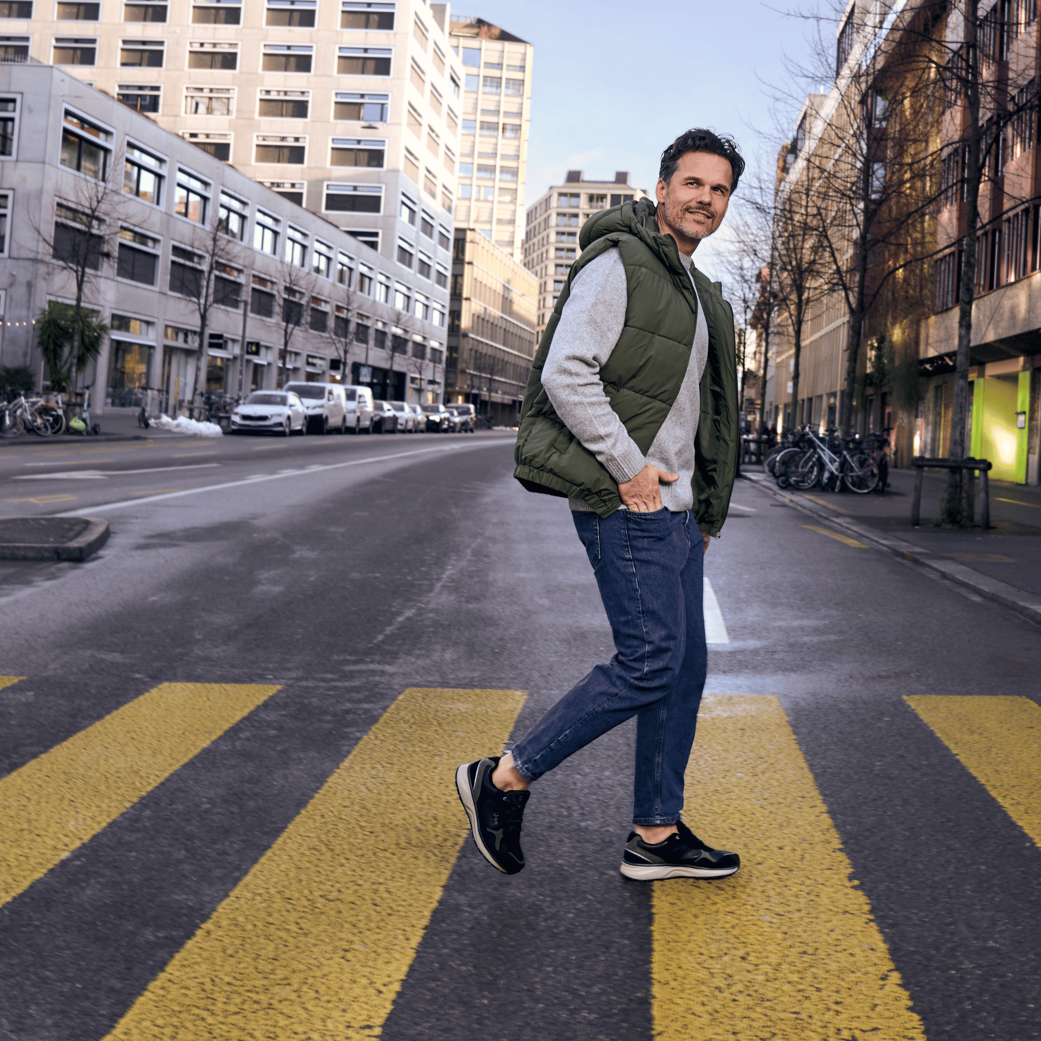 A man walking across a yellow-striped crosswalk on an urban street, wearing a green puffer vest over a light gray long-sleeve shirt, blue jeans, and black sneakers.