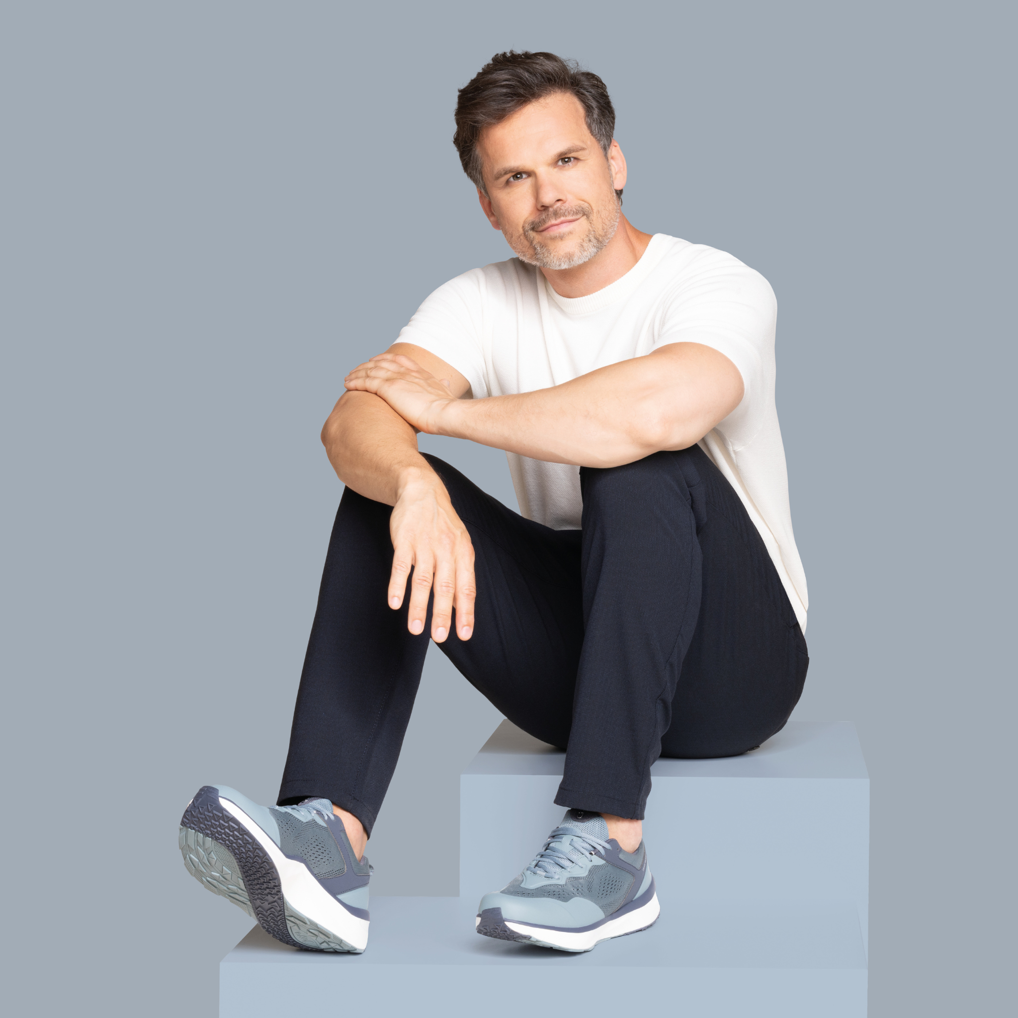 Man sitting on a block wearing a white shirt, black pants, and gray sneakers against a gray background