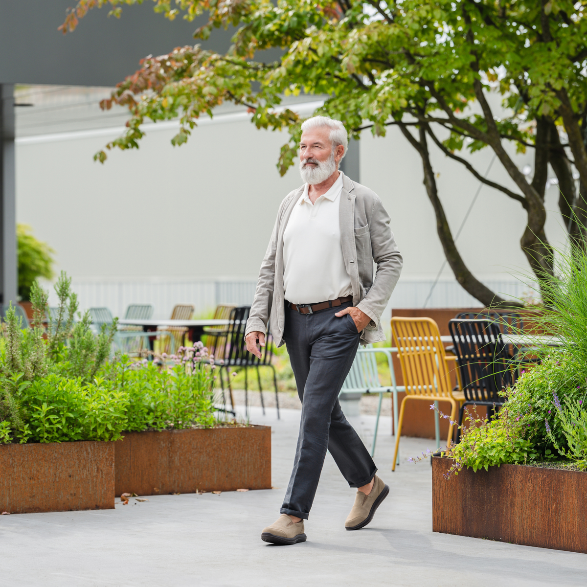 An older man with a white beard walks outdoors in a modern patio setting, wearing a light gray blazer, white polo shirt, dark gray trousers, and brown shoes.