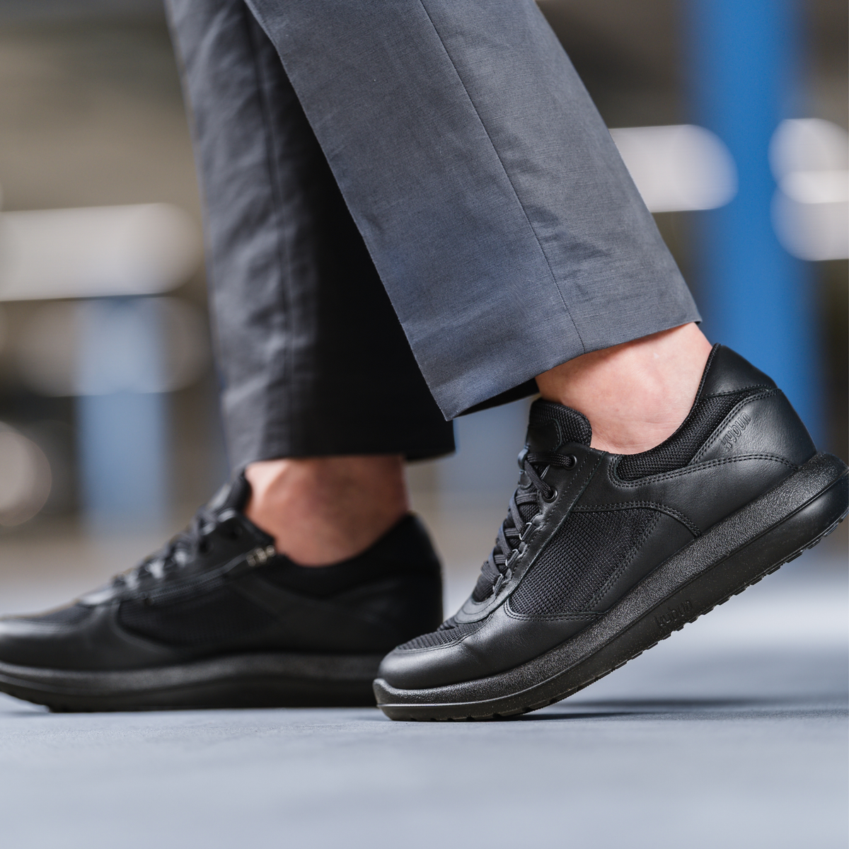 A close-up of a person wearing black low-top sneakers with gray trousers, captured in a blurred indoor setting.