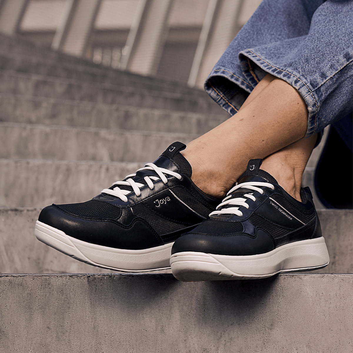 women in jeans wearing joya's black and white sneaker with grey concrete steps on the background.