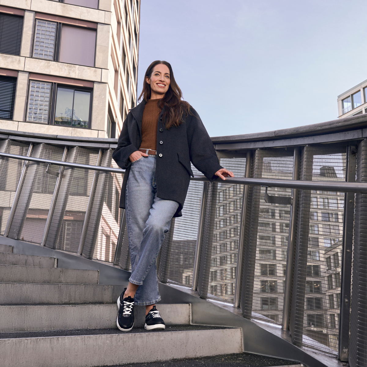 A woman stands on outdoor concrete stairs in a modern urban setting, wearing a black oversized coat over a brown top, light blue jeans, and black sneakers with white soles.