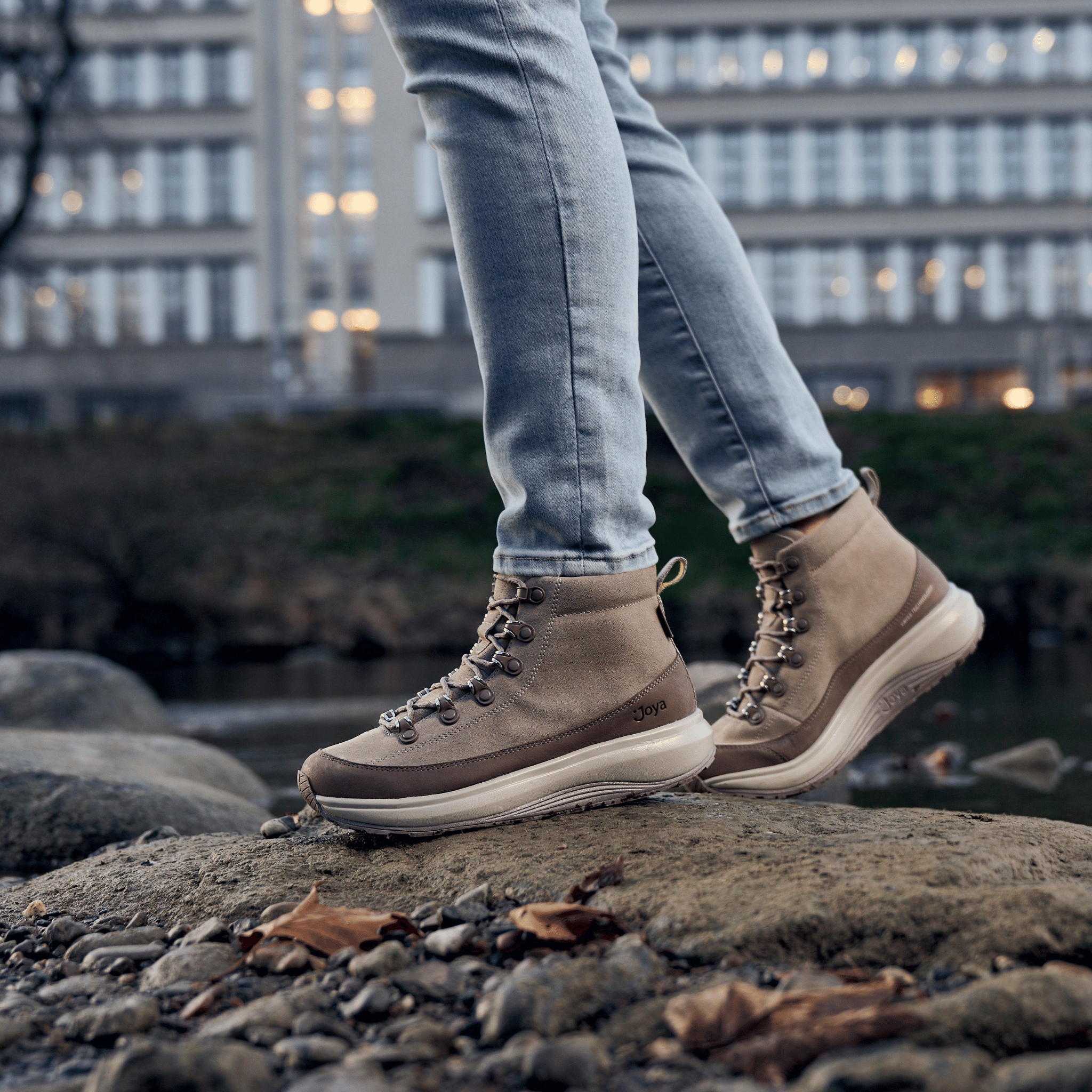 A person wearing beige and brown hiking boots with light blue jeans, standing on a rocky riverbank with a blurred city background.