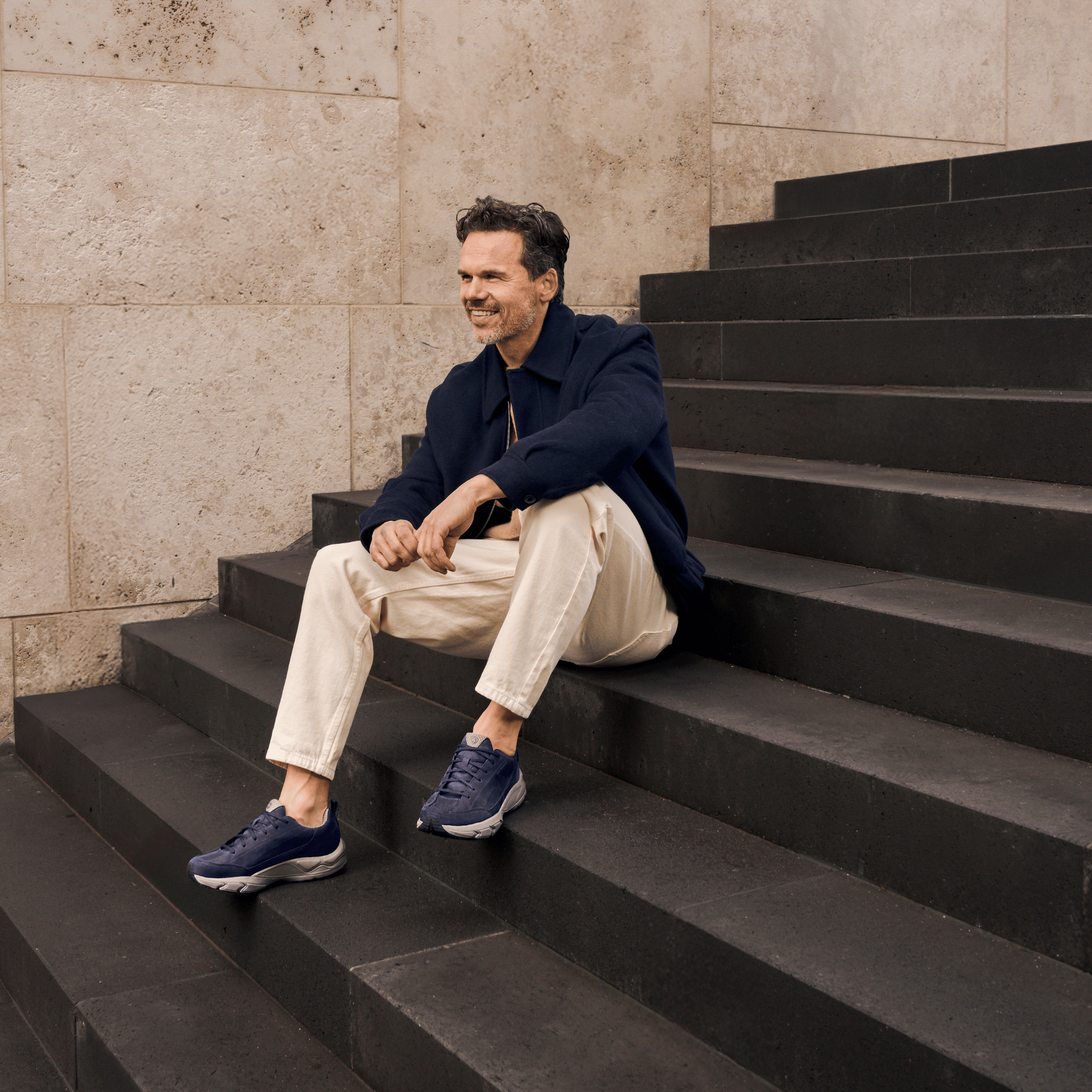 A man sits on dark stone steps wearing a navy jacket, beige trousers, and navy sneakers against a textured beige stone wall.