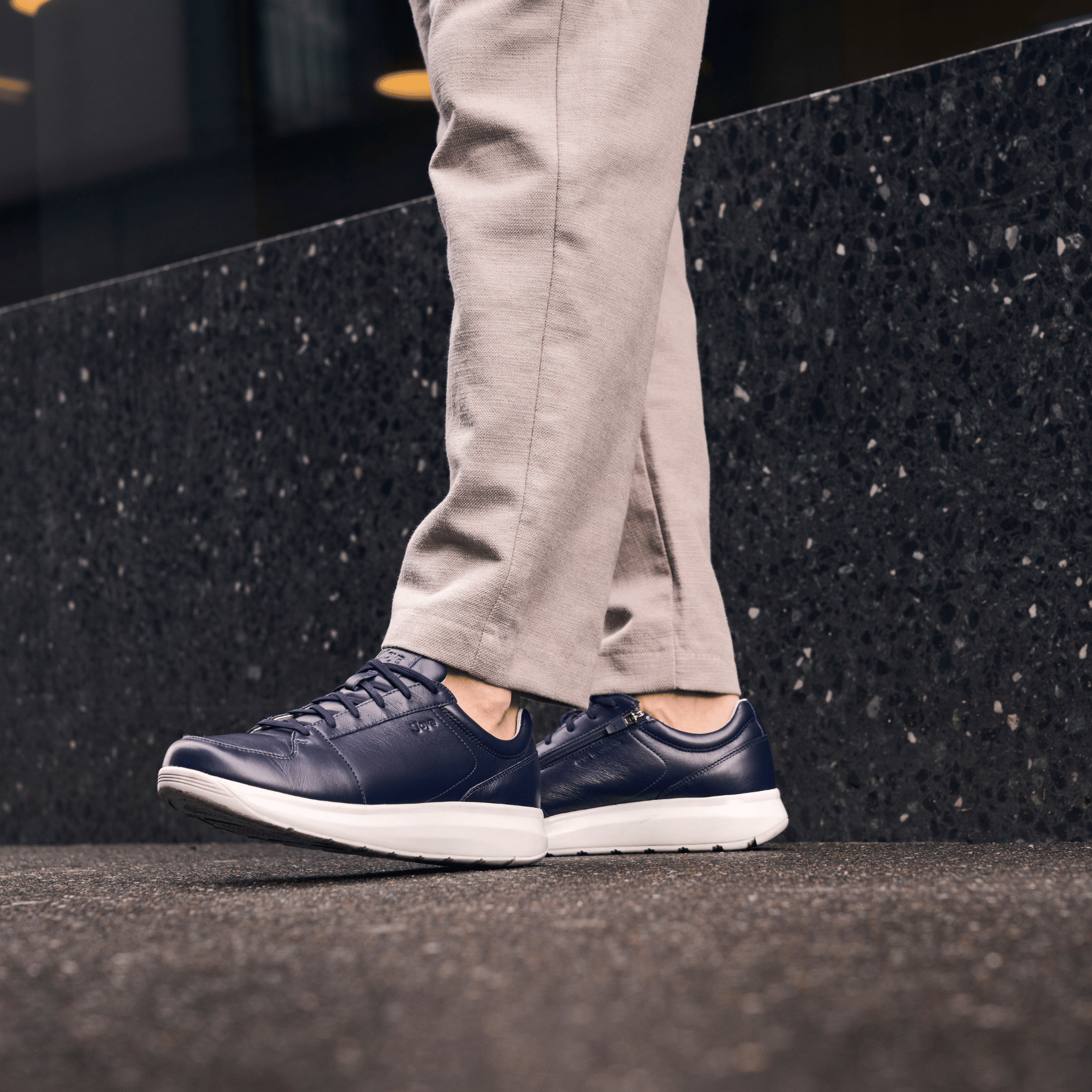 A person wearing navy blue leather low-top sneakers with white soles, paired with light beige trousers, standing on a dark textured stone ledge.