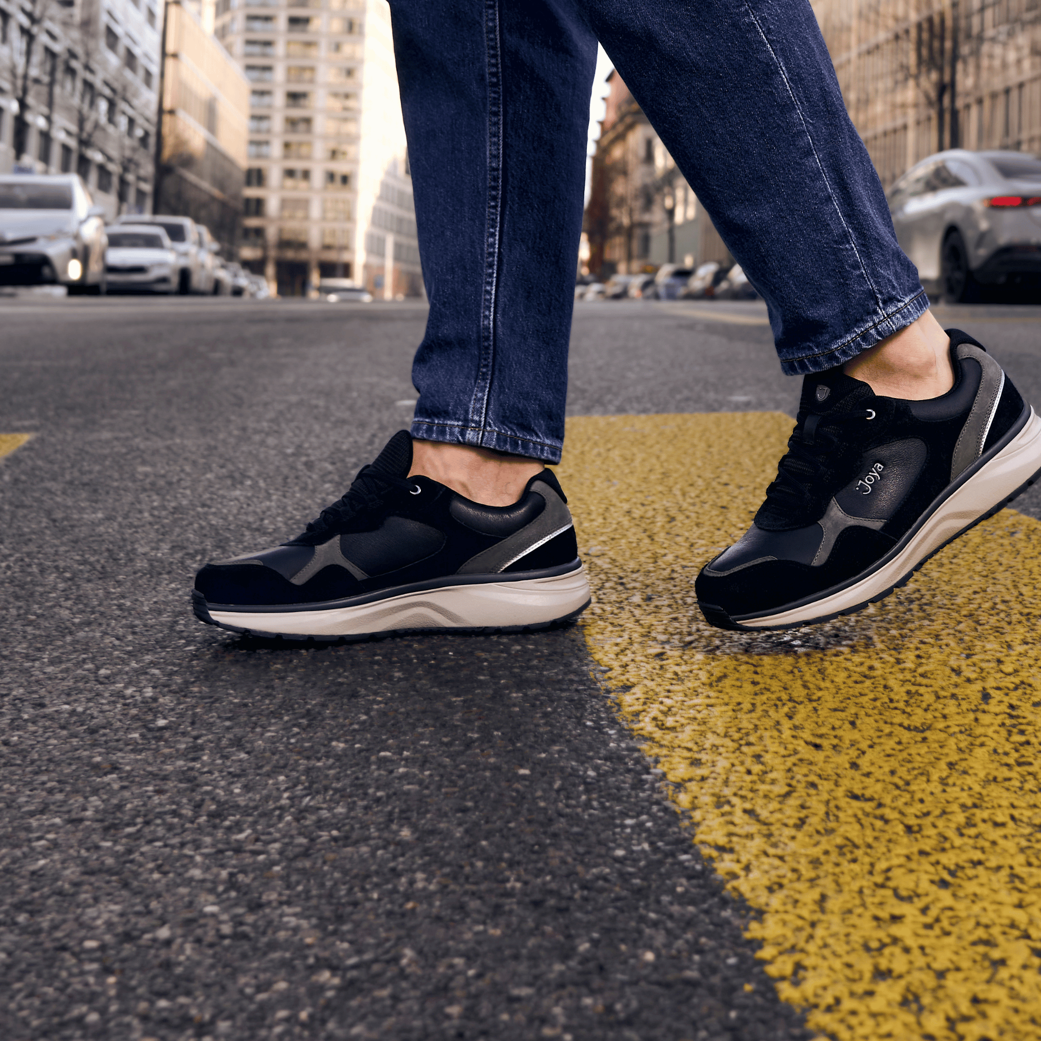 A person wearing black and gray athletic sneakers with blue jeans walking on a city street with a yellow crosswalk line.