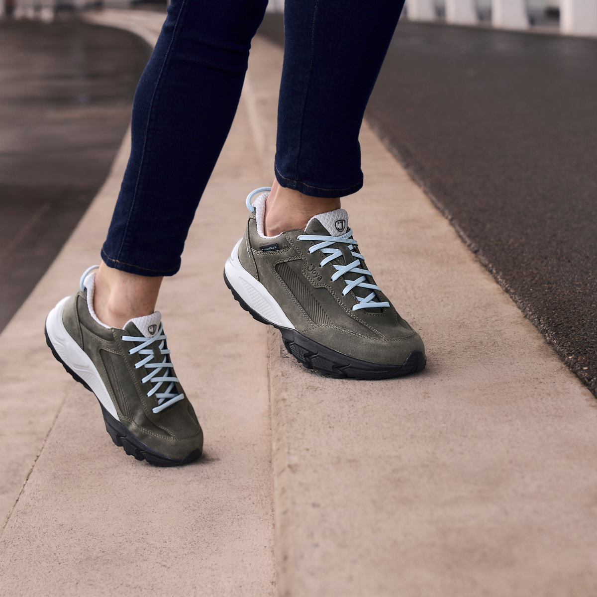 A person wearing olive green and white sneakers with light blue laces, paired with dark blue jeans, walking on a concrete path outdoors.