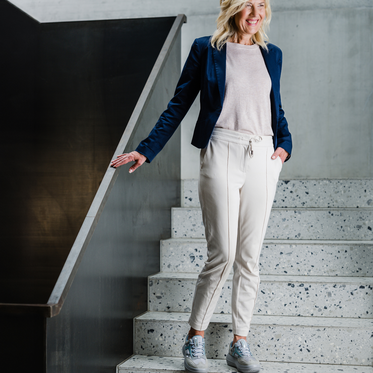 A woman stands on indoor terrazzo stairs wearing a navy blazer, light pink top, off-white drawstring trousers, and gray sneakers.