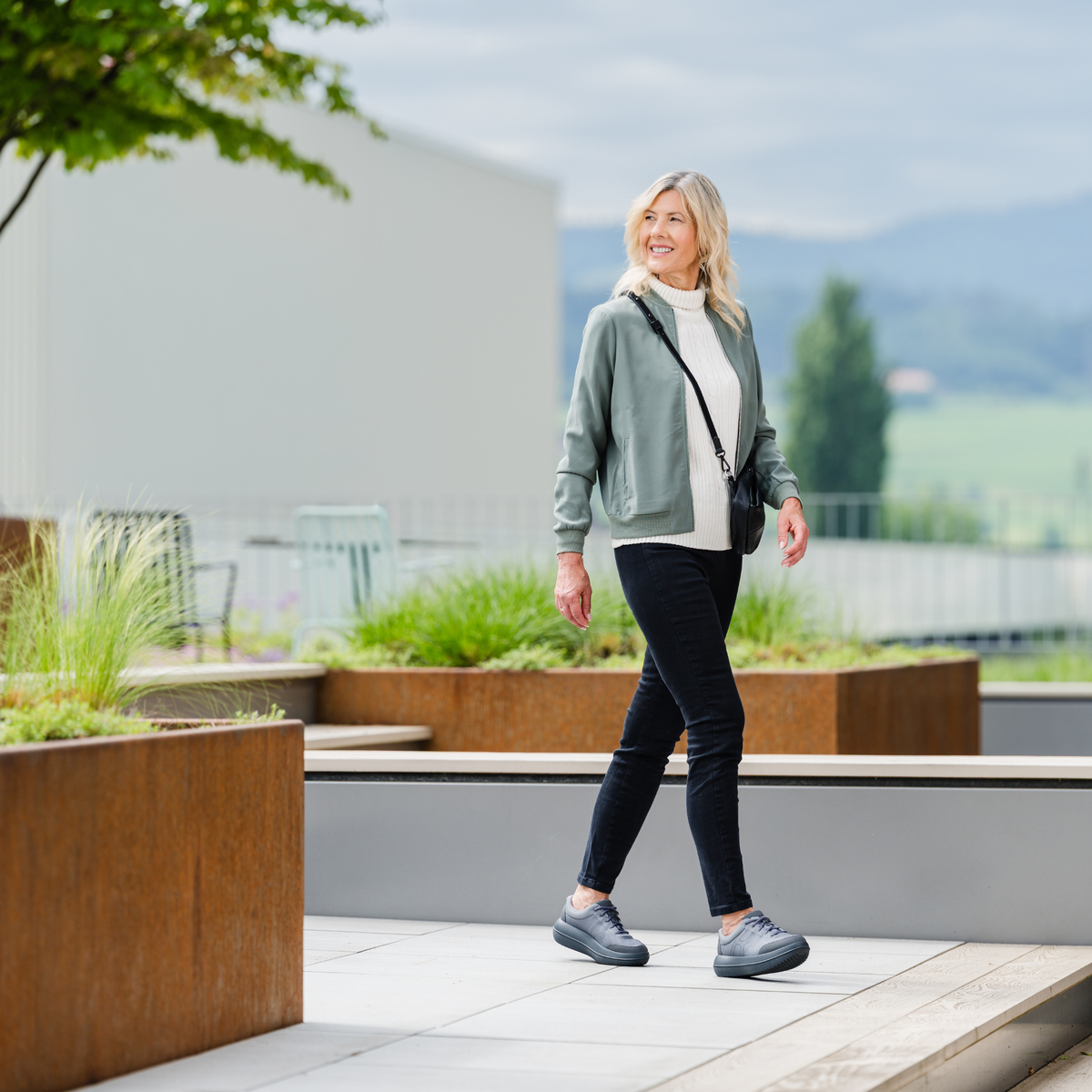 A woman walking outdoors on a modern terrace, wearing a sage green zip-up hoodie over a white turtleneck, black skinny jeans, and gray sneakers, with a black shoulder bag.