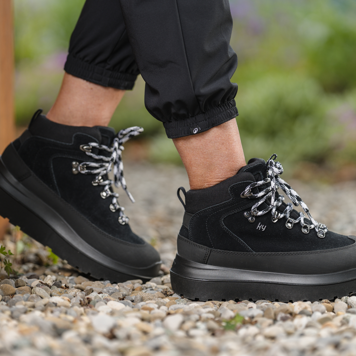 Black ankle hiking boots with chunky black soles and silver-toned metal eyelets, shown on a person wearing black pants while walking on a gravel path outdoors.