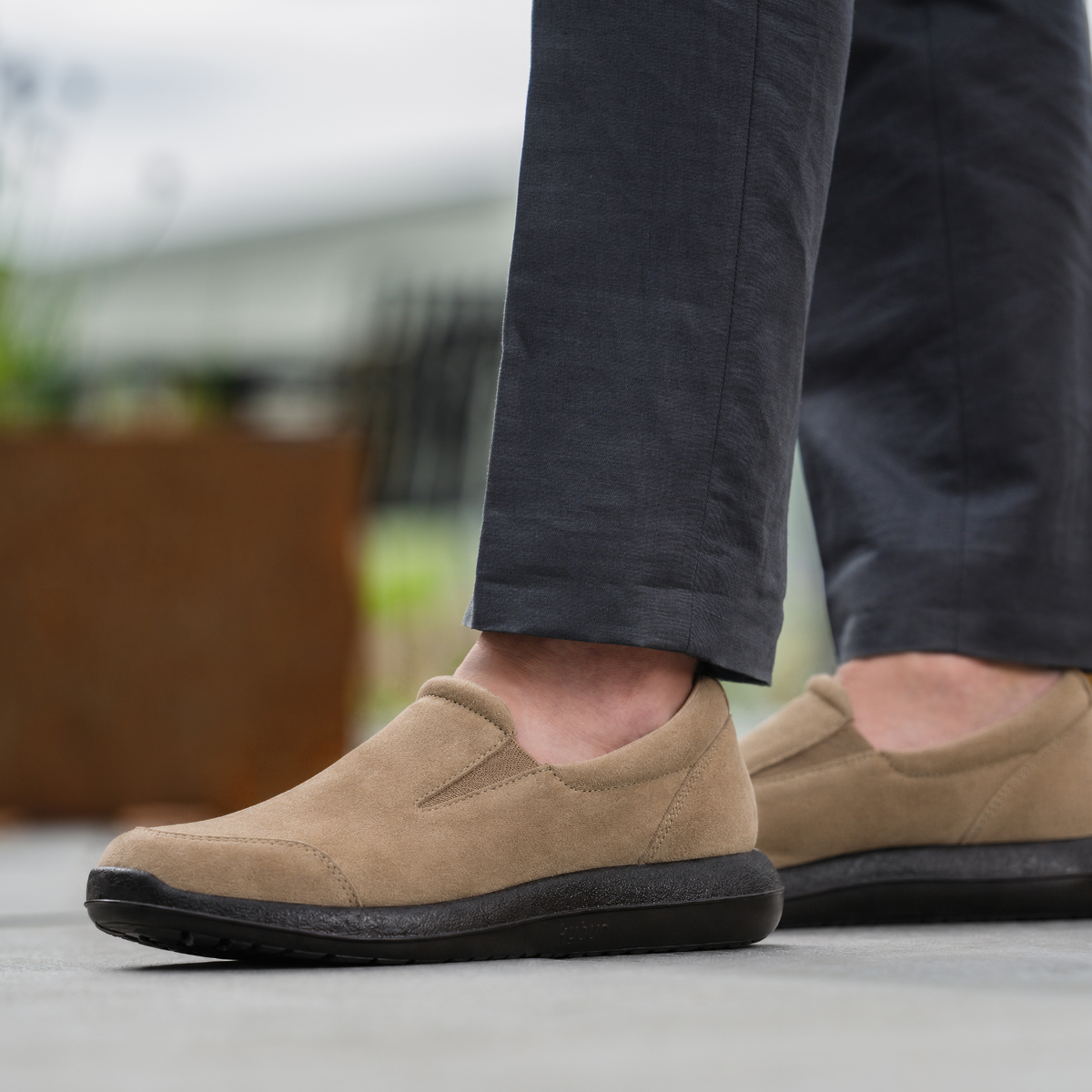 A close-up of a person's feet wearing tan slip-on shoes with dark soles, paired with dark gray trousers, standing on a concrete surface.
