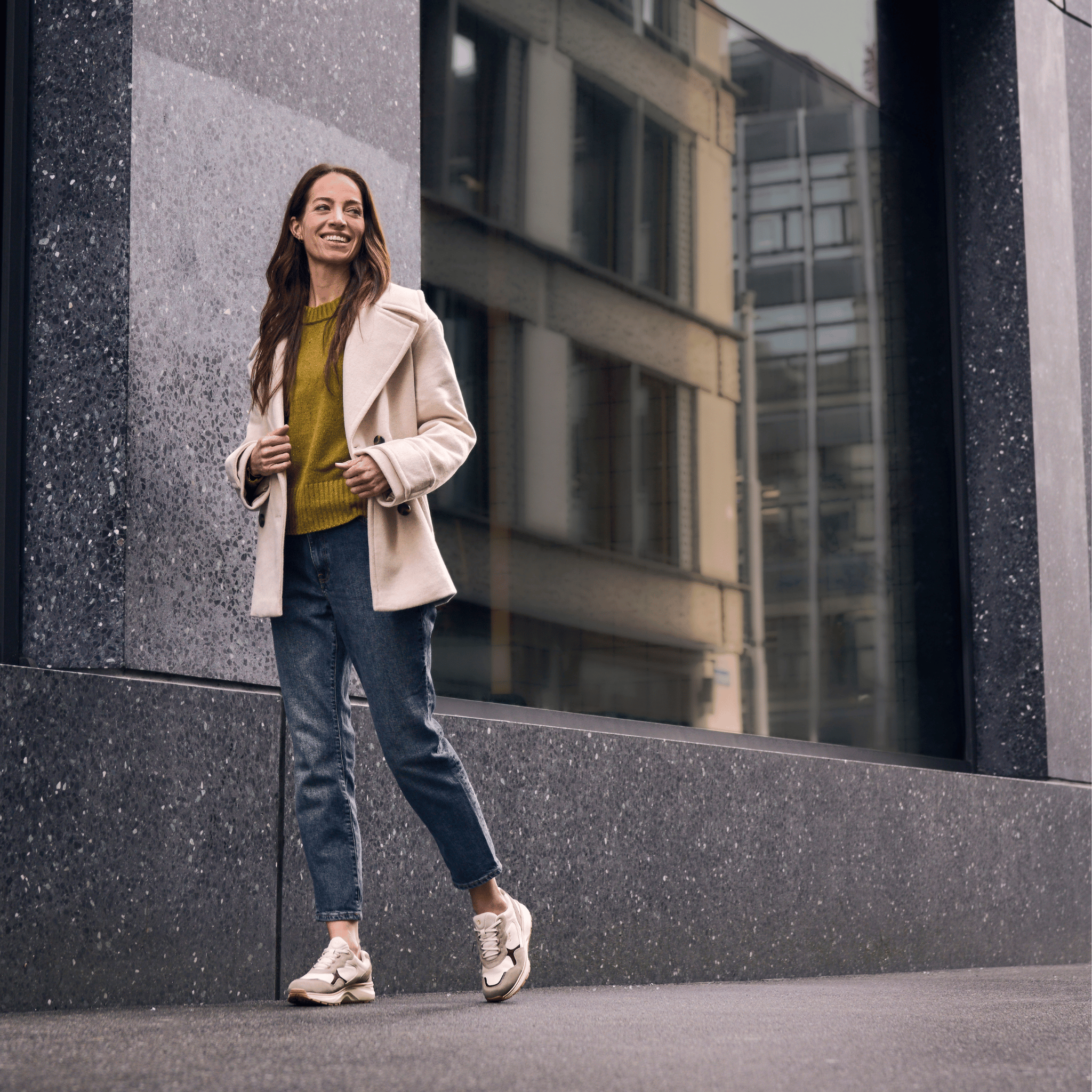 women wearing jeans, beige jacket and beige sneakers walking on the street with a blurred building in the background