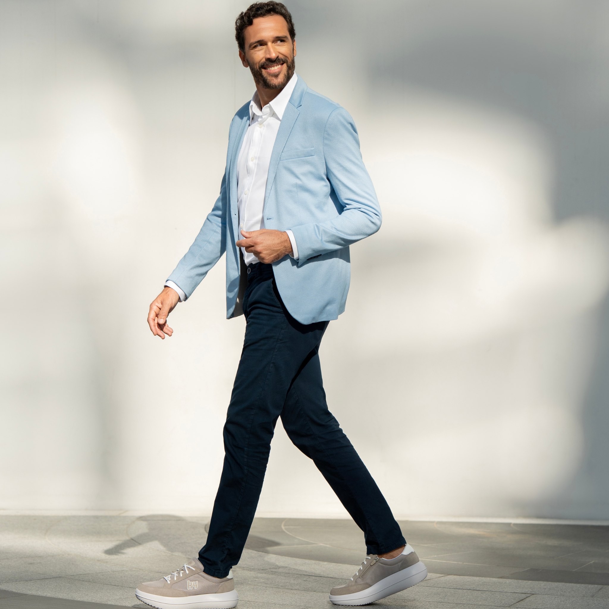 Man wearing a light blue blazer over a white shirt, dark pants, and gray shoes on a neutral background